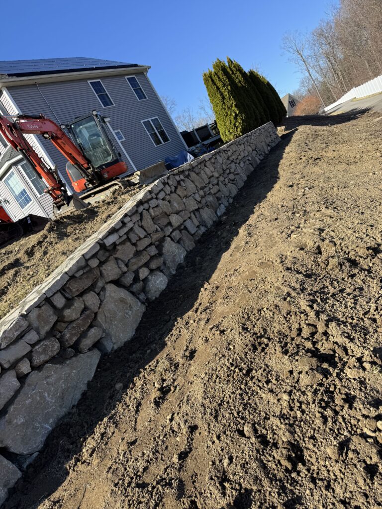 Stone retaining wall construction along sloped yard with excavator and house in Connecticut.